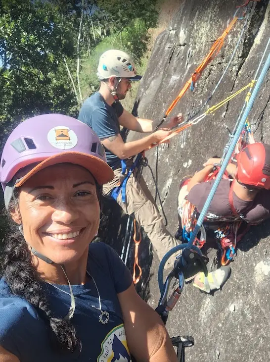 Danielle Pinto sorrindo na parede durante uma aula prática, com alunos treinando manobras de resgate ao fundo