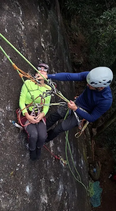 Instrutor posicionado ao lado de uma aluna suspensa, demonstrando manobra com corda verde
