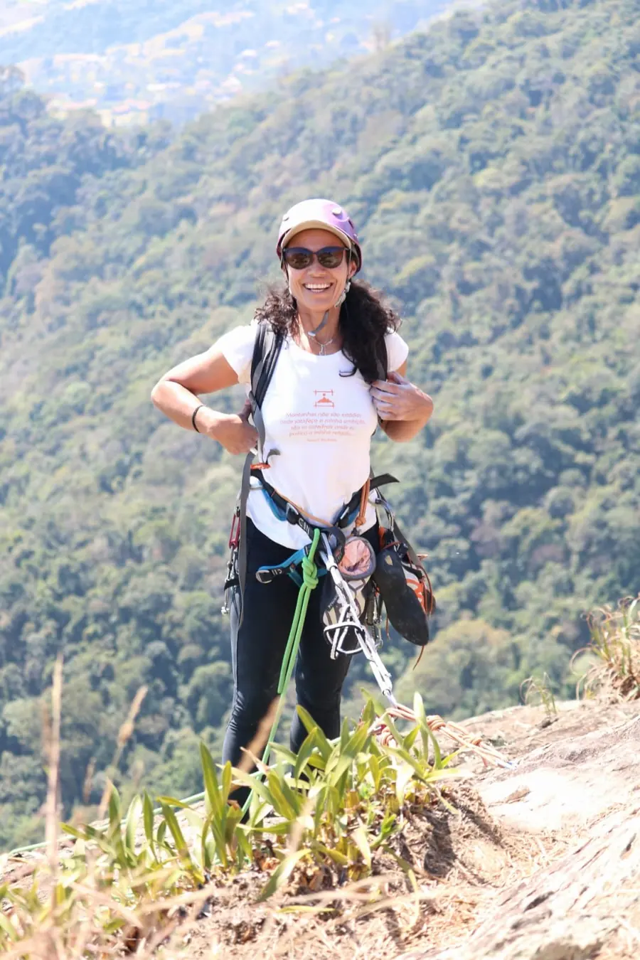 Danielle Pinto sorrindo no alto da via, com equipamento de escalada e a mata da Serra da Mantiqueira ao fundo