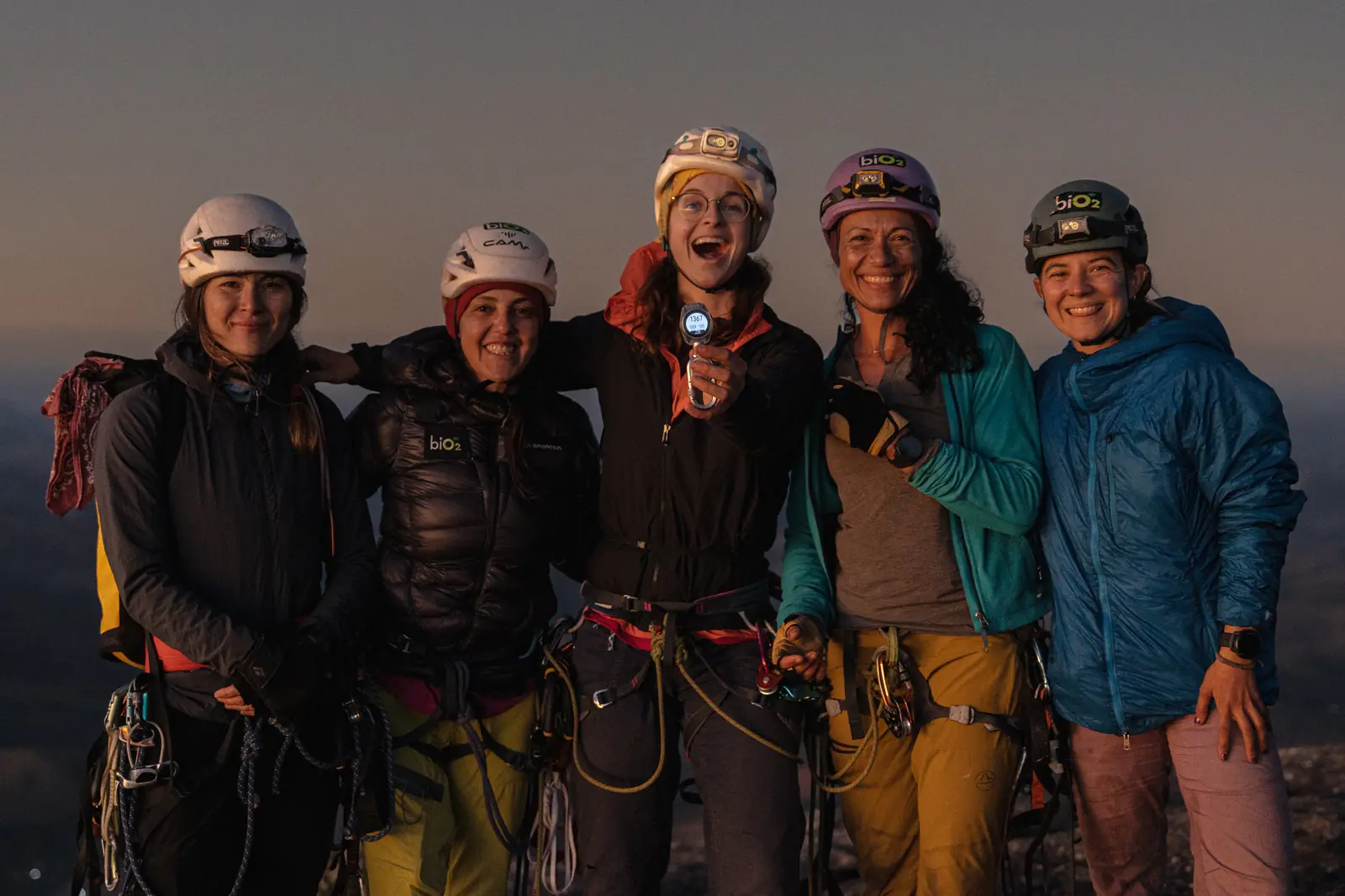 Grupo de escaladoras abraçadas e sorrindo ao entardecer, de capacete e lanterna
