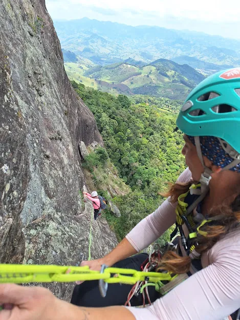 Escaladora de capacete verde fazendo segurança no alto de uma parede, com o vale ao fundo