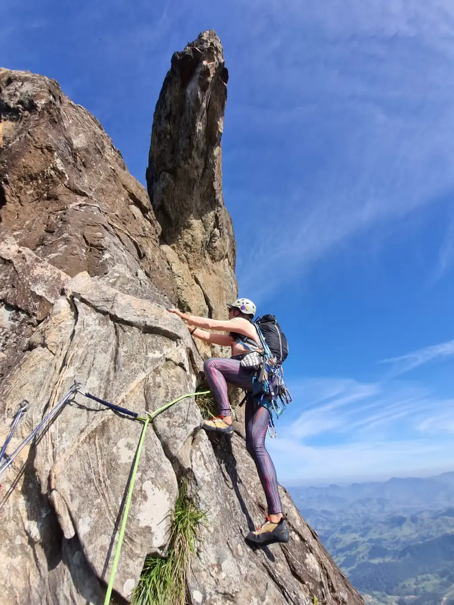 Escaladora de capacete branco subindo a rocha ao lado de uma agulha de pedra, com o vale ao fundo