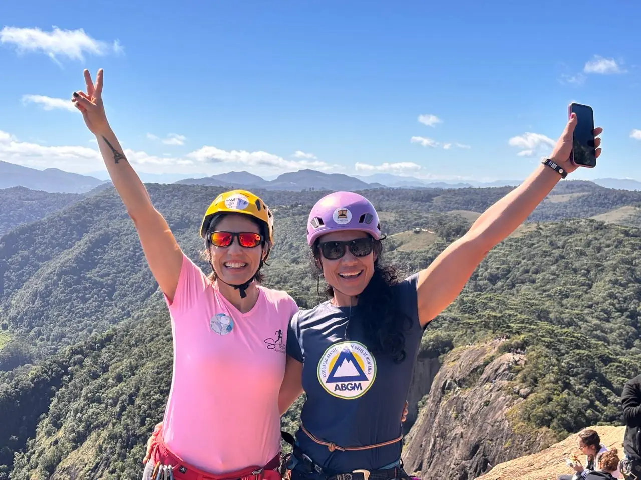 Danielle Pinto e uma escaladora comemorando no cume, de braços para cima, com a Serra da Mantiqueira ao fundo
