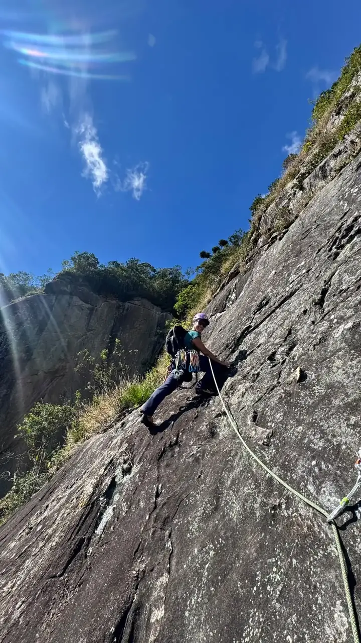 Escaladora sorrindo em uma placa de granito sob céu azul, guiando a corda verde