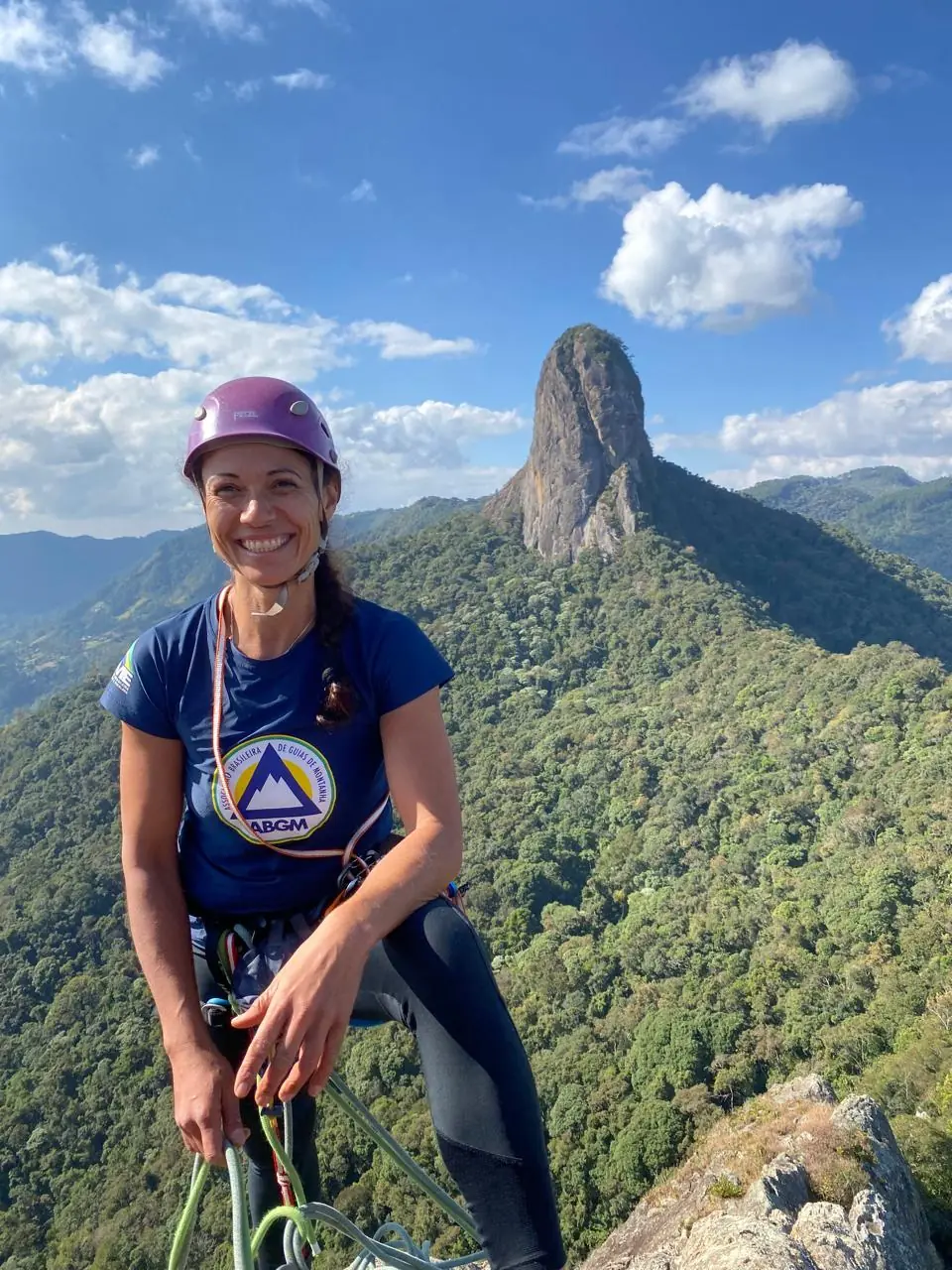 Danielle Pinto sorrindo no cume, de capacete e camiseta da ABGM, com o Bauzinho ao fundo