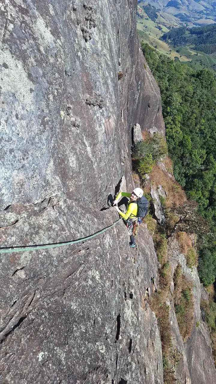 Escalador de capacete branco e camiseta amarela subindo uma grande parede de granito, com a mata lá embaixo