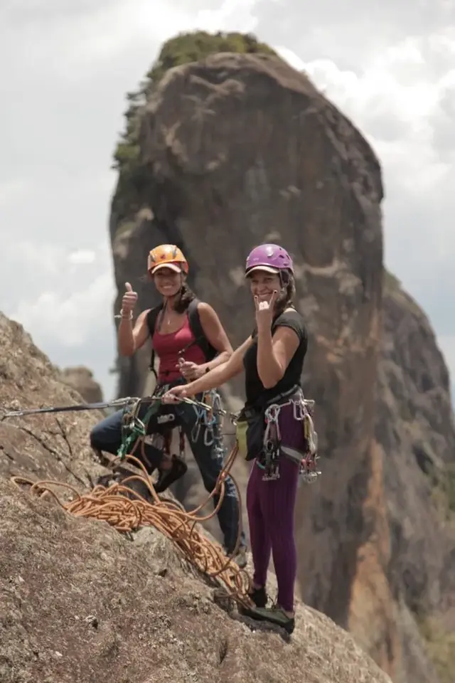 Duas escaladoras sorrindo de capacete com uma grande pedra ao fundo