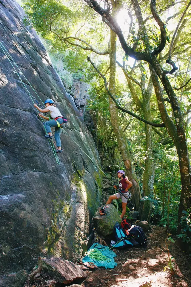 Aluna escalando uma parede de rocha na mata, com a instrutora fazendo a segurança