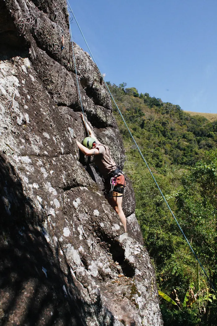 Escalador de capacete verde subindo a parede, com a mata da serra ao fundo