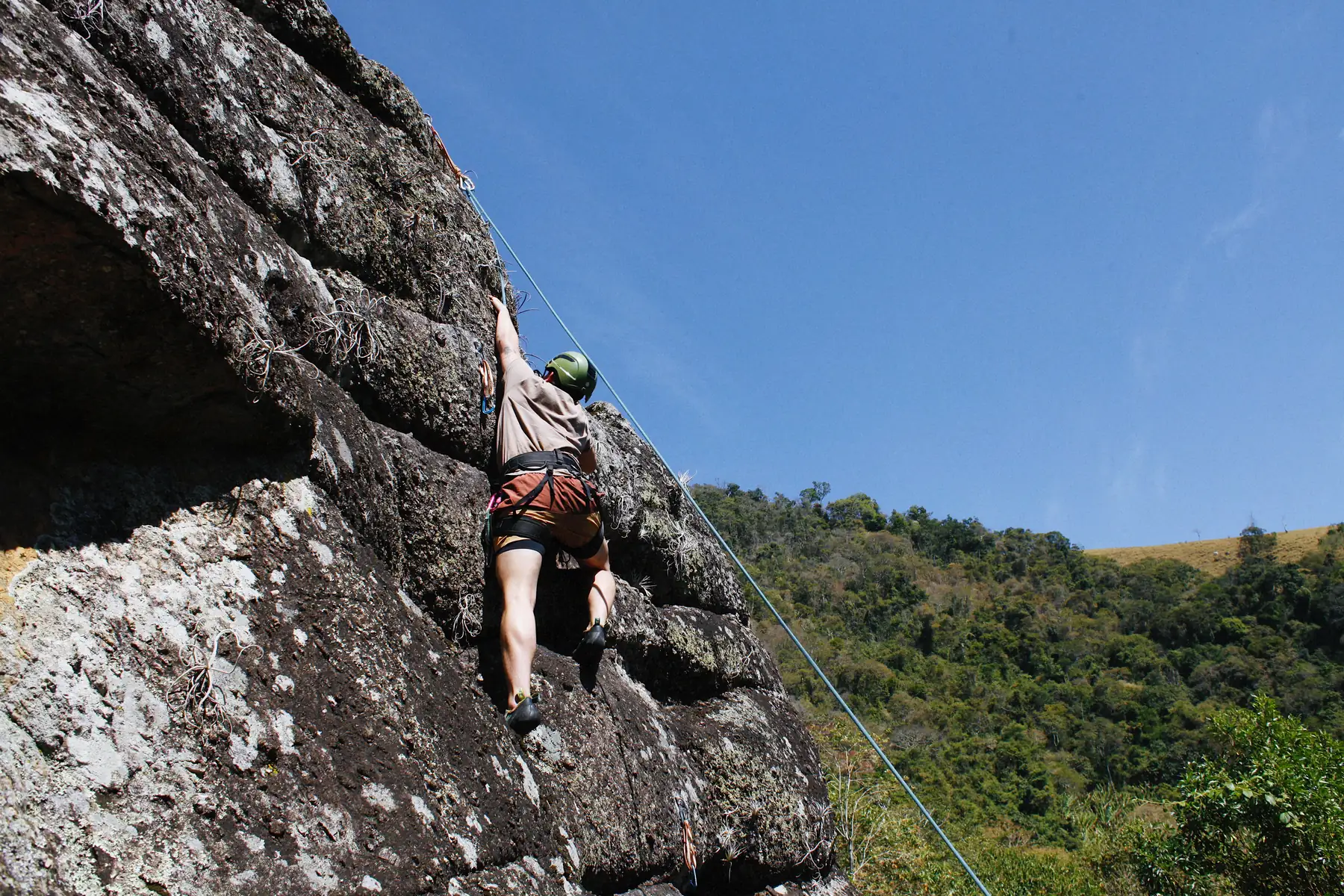 Escalador de capacete verde subindo a parede de rocha sob céu azul, com a mata da Serra da Mantiqueira ao fundo