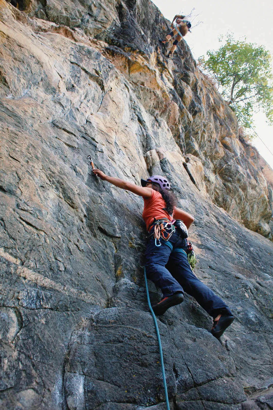 Escaladora de capacete lilás clipando a costura na parede de rocha, com a instrutora acompanhando do alto
